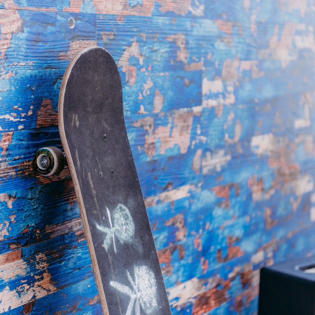 Ein gebrauchtes Skateboard lehnt an einer Wandverkleidung aus Holzpaneelen in einem intensiven, verwaschenen Blauton mit Vintage-Effekten. Die Kombination aus Urban-Style und rustikalem Holzdekor erzeugt einen individuellen und modernen Look.