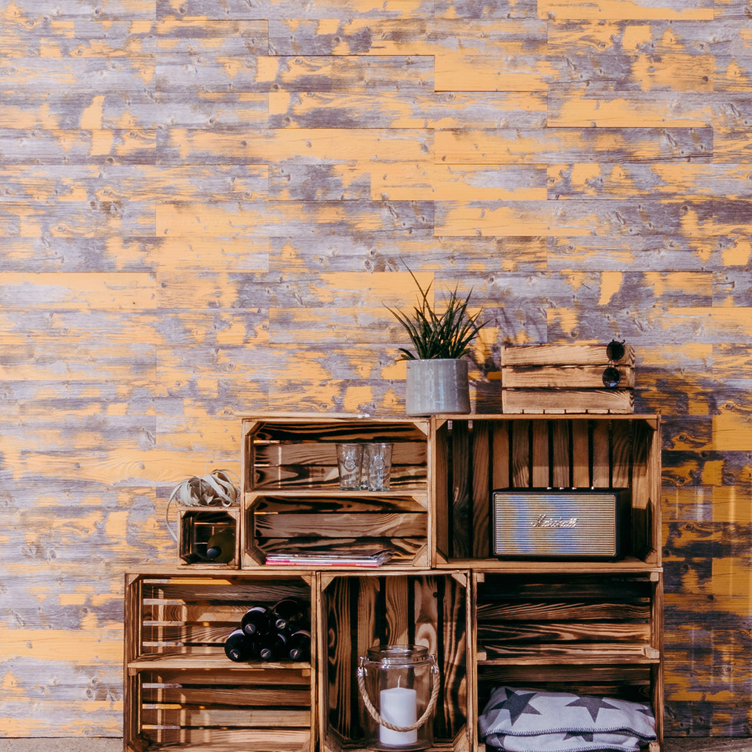 Ein kreatives Regalsystem aus geflammten Obstkisten ist vor einer auffälligen Wand in gelb-blauer Vintage-Holzoptik aufgebaut. Die Kisten dienen als dekorative Ablage für Weinflaschen, Gläser und eine Musikbox im Retro-Stil.