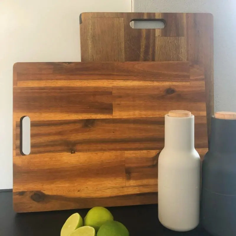 Zwei glatte Servierbretter aus Holz lehnen hochkant an einer Wand, um nach der Reinigung zu trocknen. Davor stehen zwei Salz- und Pfeffermühlen sowie einige frische Limetten.