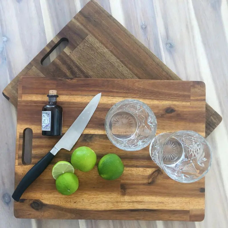 Zwei gepflegte Holz-Schneidebretter liegen versetzt übereinander. Auf dem oberen Brett sind frische Limetten, ein Küchenmesser, zwei Gläser und ein kleines Fläschchen arrangiert.