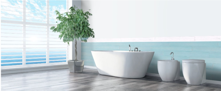 Ein luxuriöses Badezimmer mit einer freistehenden weißen Badewanne und passenden Sanitärelementen vor einer halbhoch verkleideten Wand in hellblauem Holzdekor. Das lichtdurchflutete Zimmer bietet durch ein großes Fenster mit Jalousien einen Blick auf das Meer.