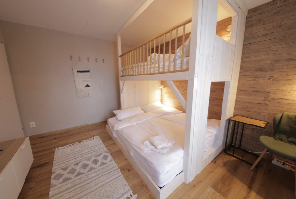 Ein modernes, weiß lasiertes Etagenbett aus massivem Holz in einem hellen Zimmer mit Echtholzboden. Das Bett ist vor einer Wand mit rustikalen Holzpaneelen platziert, was zusammen mit einem kleinen Teppich und einem grünen Sessel eine behagliche Wohnatmosphäre schafft.