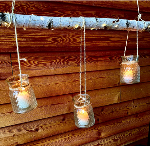 Drei gläserne Windlichter mit brennenden Kerzen hängen an Seilen von einem horizontalen Birkenast herab. Eine feine Lichterkette umwickelt den Ast und sorgt für eine stimmungsvolle Beleuchtung vor einer rötlichen Holzwand.