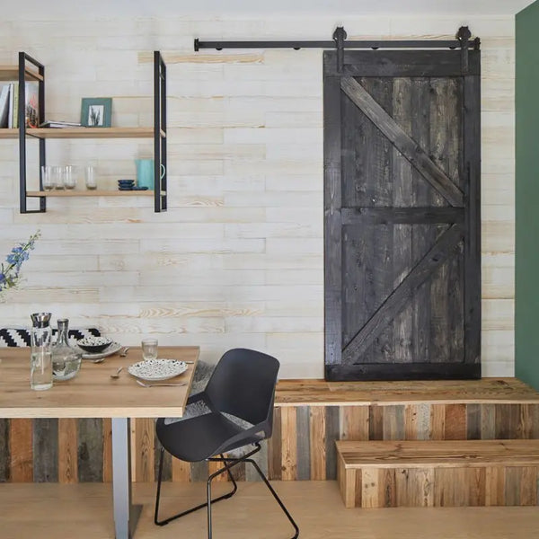 Ein modern eingerichtetes Esszimmer mit einer hellen Holzpaneel-Wand, an der eine dunkle, massive Schiebetür angebracht ist. Ein Podest im Hintergrund ist als rustikaler Kontrast mit bunten Altholzpaneelen verkleidet.