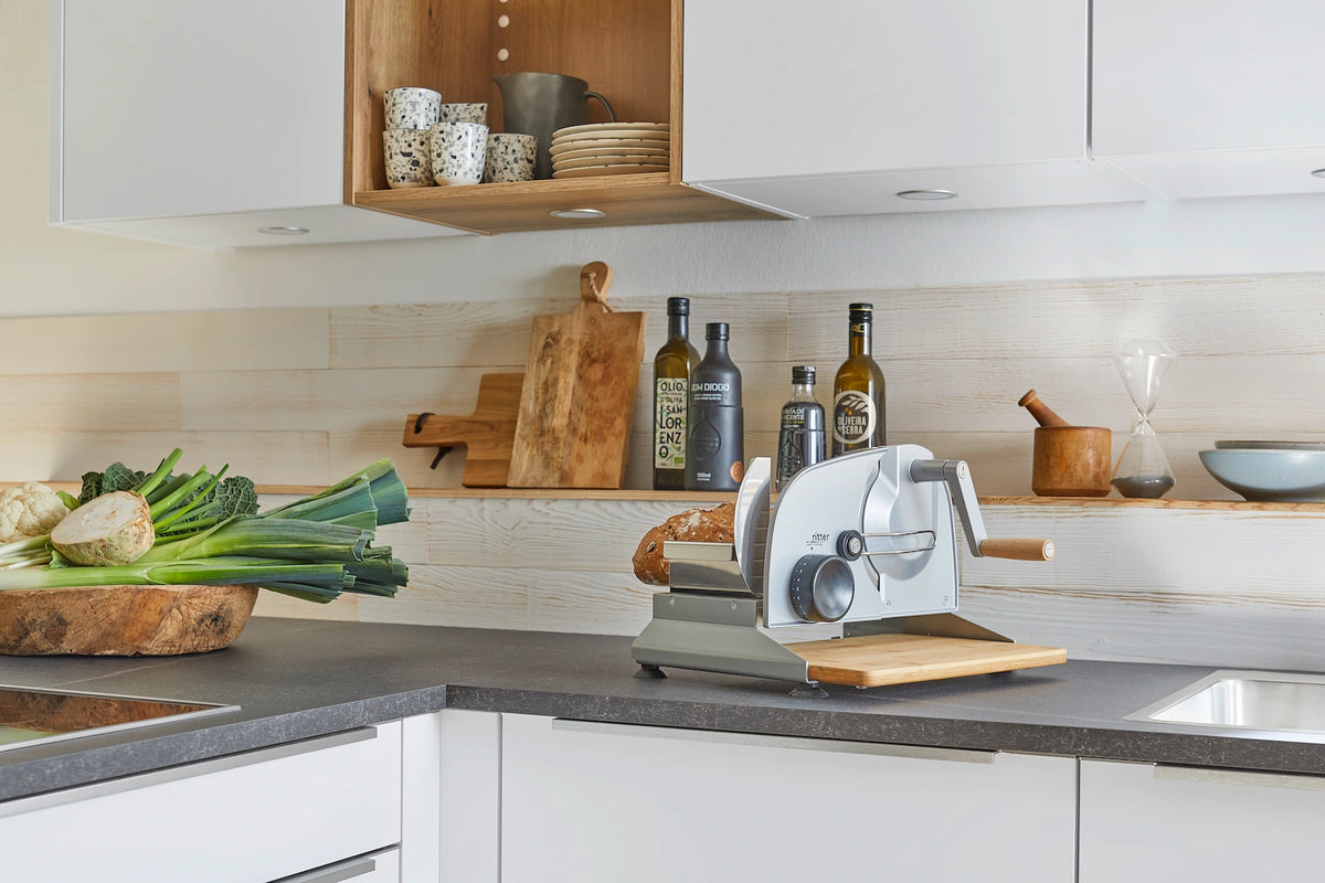 Eine moderne weiße Küche mit dunkler Arbeitsplatte, die durch helle Holzpaneele ergänzt wird. Auf der Arbeitsfläche stehen frisches Gemüse und eine klassische Brotschneidemaschine, was für eine funktionale und natürliche Atmosphäre sorgt.