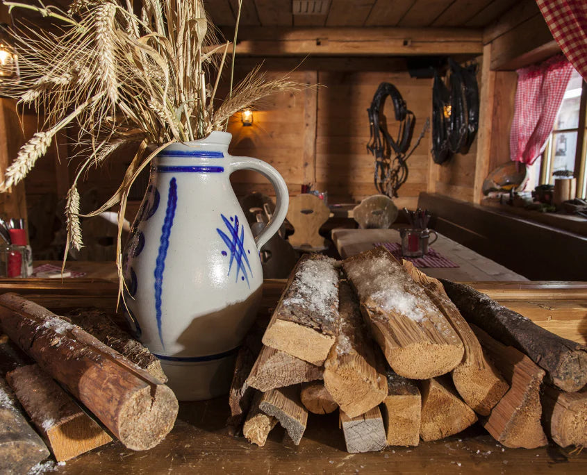 Ein traditioneller, grau-blauer Steingutkrug mit Getreideähren steht neben ordentlich aufgeschichtetem Brennholz in einer urigen Holzhütte. Die Szenerie vermittelt eine warme, ländliche Atmosphäre vor dem Hintergrund einer rustikalen Gaststube.