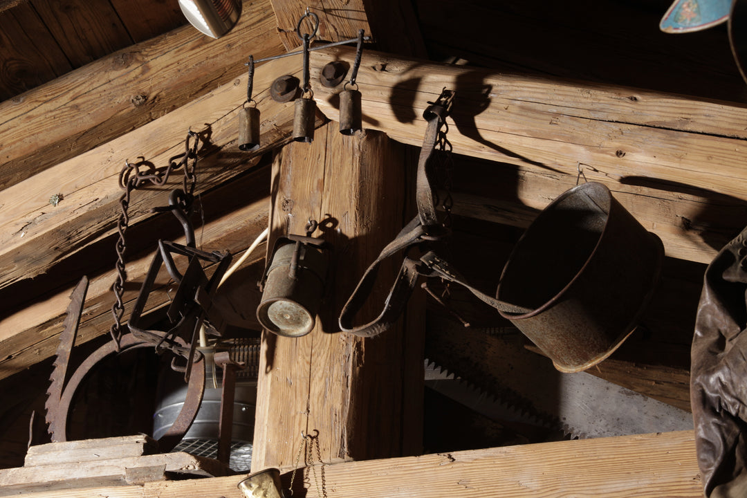 Eine Detailaufnahme verschiedener antiker landwirtschaftlicher Werkzeuge und Metallgefäße, die an einer massiven, alten Holzkonstruktion aufgehängt sind. Die Patina der Gegenstände und das grobe Holz vermitteln ein Gefühl von traditionellem Handwerk.
