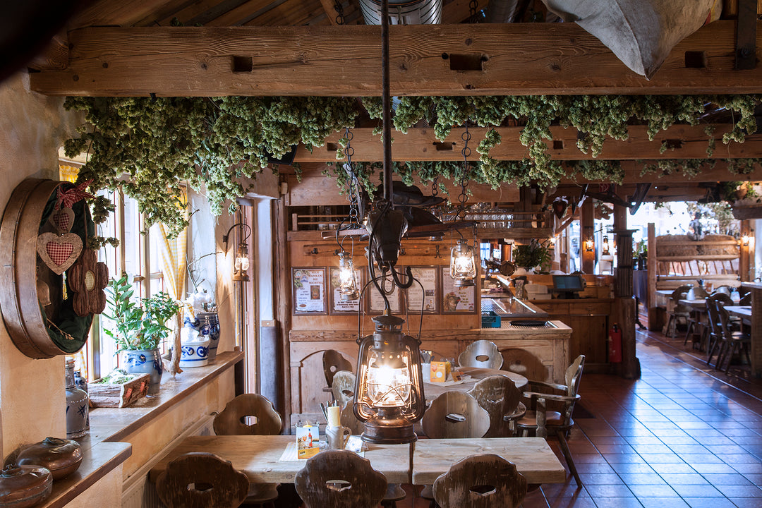 Blick in einen gemütlichen Gastraum mit schweren Holztischen und traditionell geschnitzten Stühlen. Nostalgische Petroleumlampen spenden warmes Licht, während die Dekoration aus Hopfen und ländlichen Accessoires den rustikalen Charme der Einrichtung unterstreicht.