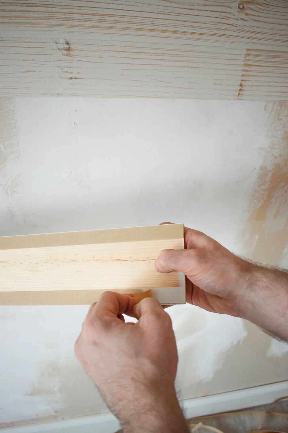 Eine Nahaufnahme zeigt zwei Hände, die die Schutzfolie von einem selbstklebenden Holzpaneel abziehen, um es an einer vorbereiteten Wand zu befestigen.