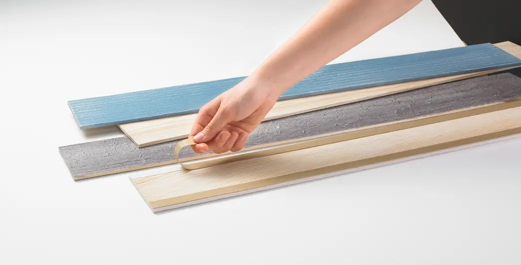 Eine Hand zieht den Klebeschutz von einem hellen Holzpaneel ab, das zusammen mit blauen und grauen Paneelen auf einer weißen Fläche liegt. Die Auswahl an unterschiedlichen Farben und Texturen veranschaulicht die Gestaltungsmöglichkeiten der Wandverkleidung.