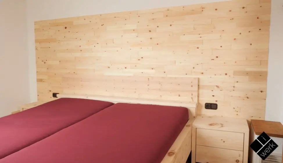 Ein massives Doppelbett aus hellem Echtholz mit passenden Nachttischen vor einer großflächigen Wandverkleidung aus natürlichen Holzpaneelen. Die durchgehende Holzoptik von Bett und Wand schafft ein warmes, harmonisches Ambiente im Schlafbereich.