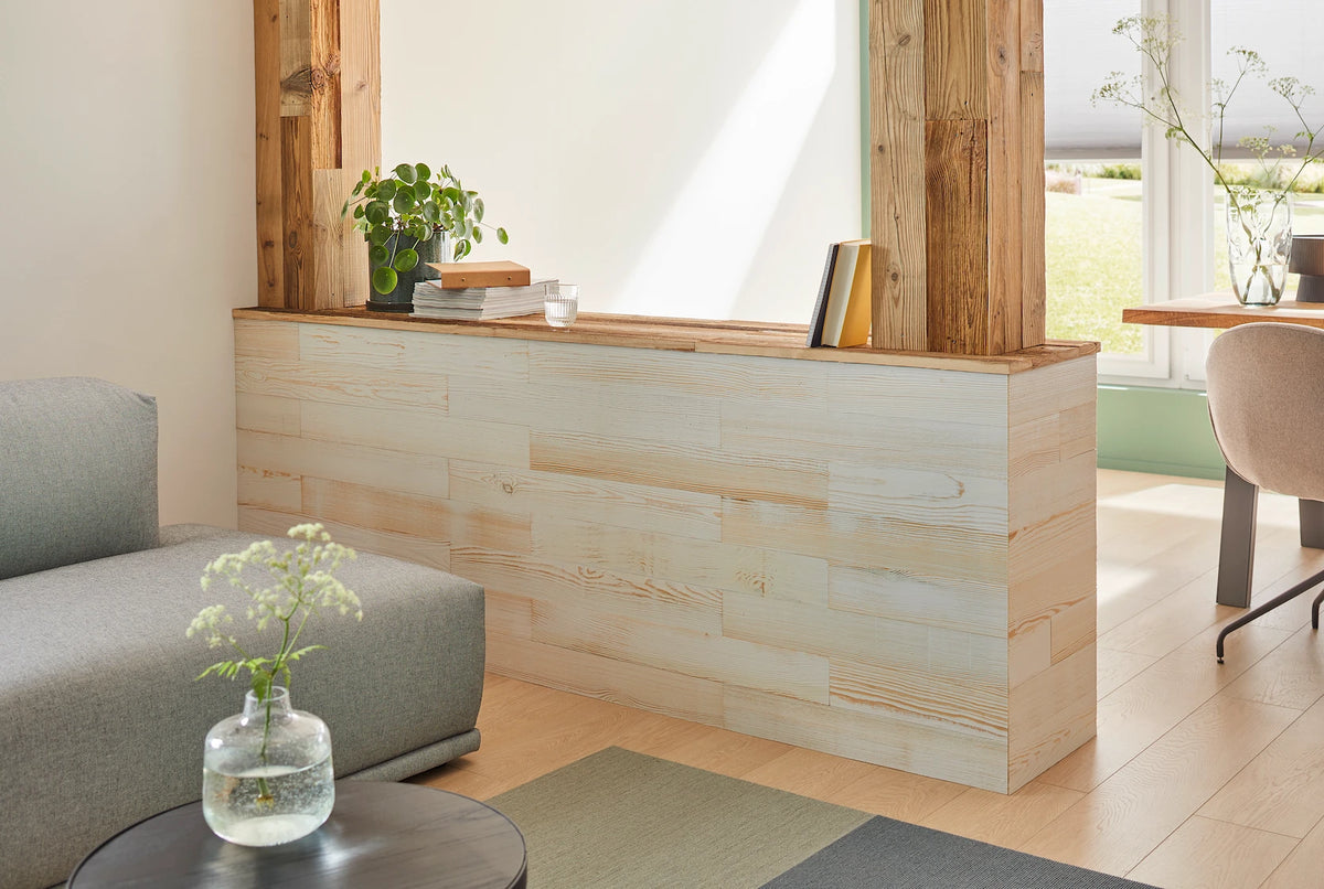 Ein halbhoher Raumteiler, der mit hellen Holzpaneelen verkleidet wurde und als Ablage für Bücher und Pflanzen dient. Zwei massive Holzsäulen rahmen das Element ein und schaffen eine natürliche Verbindung zwischen dem Wohnbereich und dem angrenzenden Esszimmer.