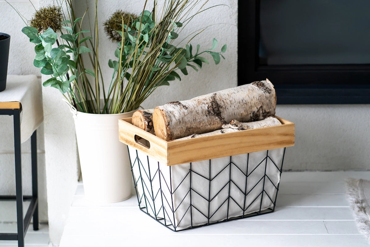 Ein moderner Drahtkorb mit Stoffeinsatz und Holzgriff ist mit zwei massiven Birkenholzscheiten gefüllt. Der Korb steht neben einer weißen Vase mit grünen Zweigen vor einem hellen Hintergrund und dient als dekorative Kaminholz-Lagerung.