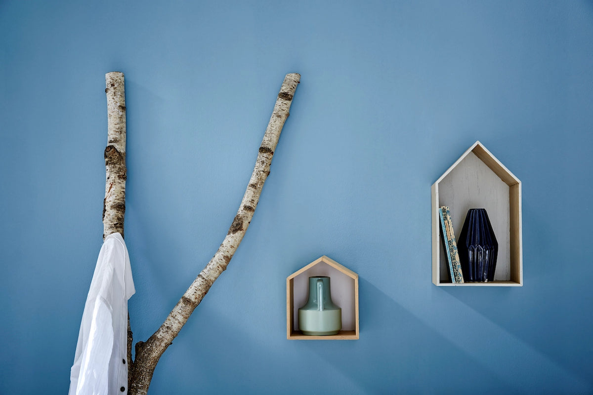 Ein verzweigter Birkenast ist als natürliches Dekorelement an einer blauen Wand befestigt und wird als Garderobenhaken genutzt. Daneben hängen zwei hausförmige Holzregale, die dezent mit Vasen und Büchern bestückt sind.