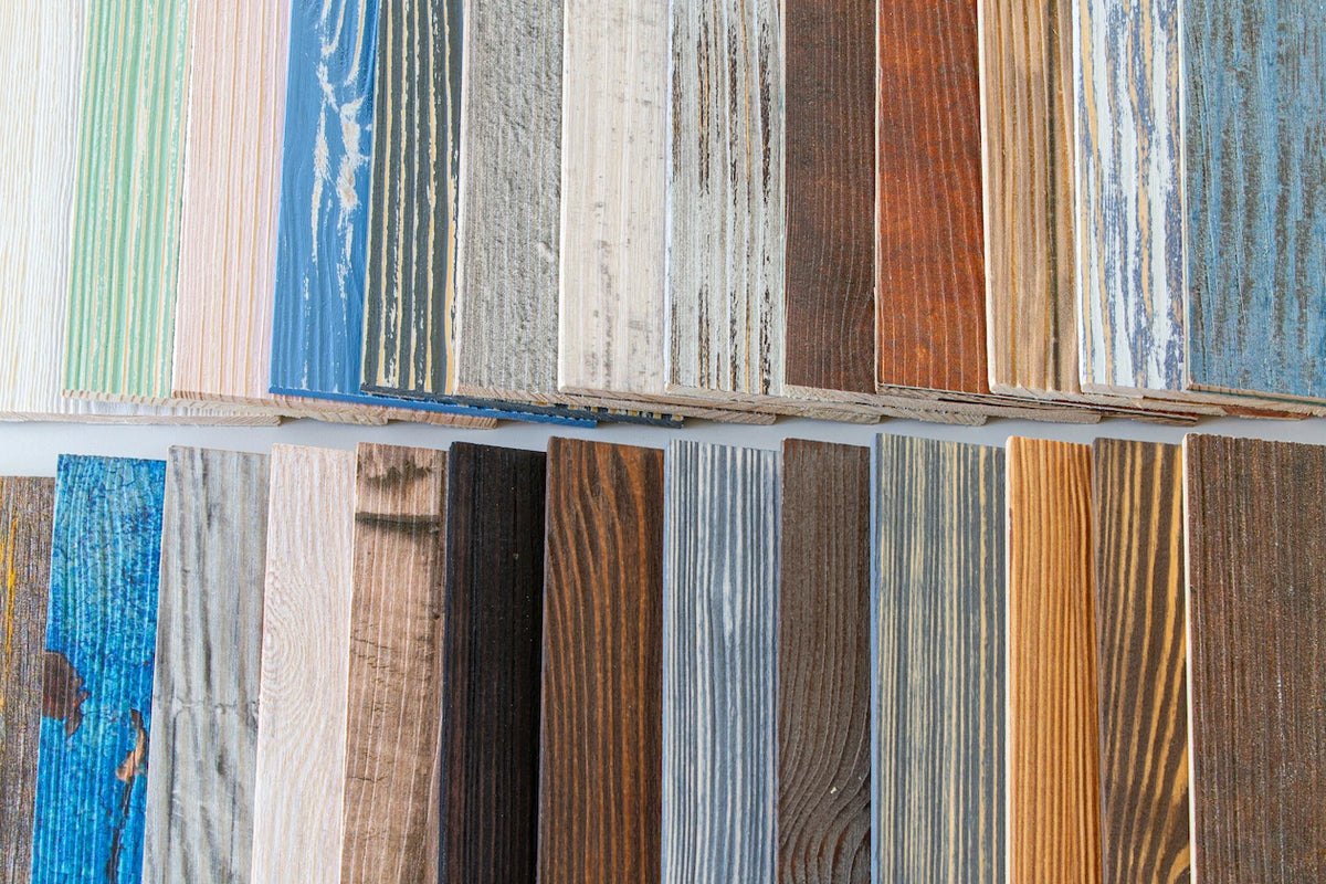 Zwei übereinanderliegende Reihen von Holzpaneel-Mustern demonstrieren die große Vielfalt an verfügbaren Farben und Oberflächenstrukturen. Die Auswahl reicht von natürlichen Holztönen über rustikale Graunuancen bis hin zu farbigen Akzenten in Blau und Grün.