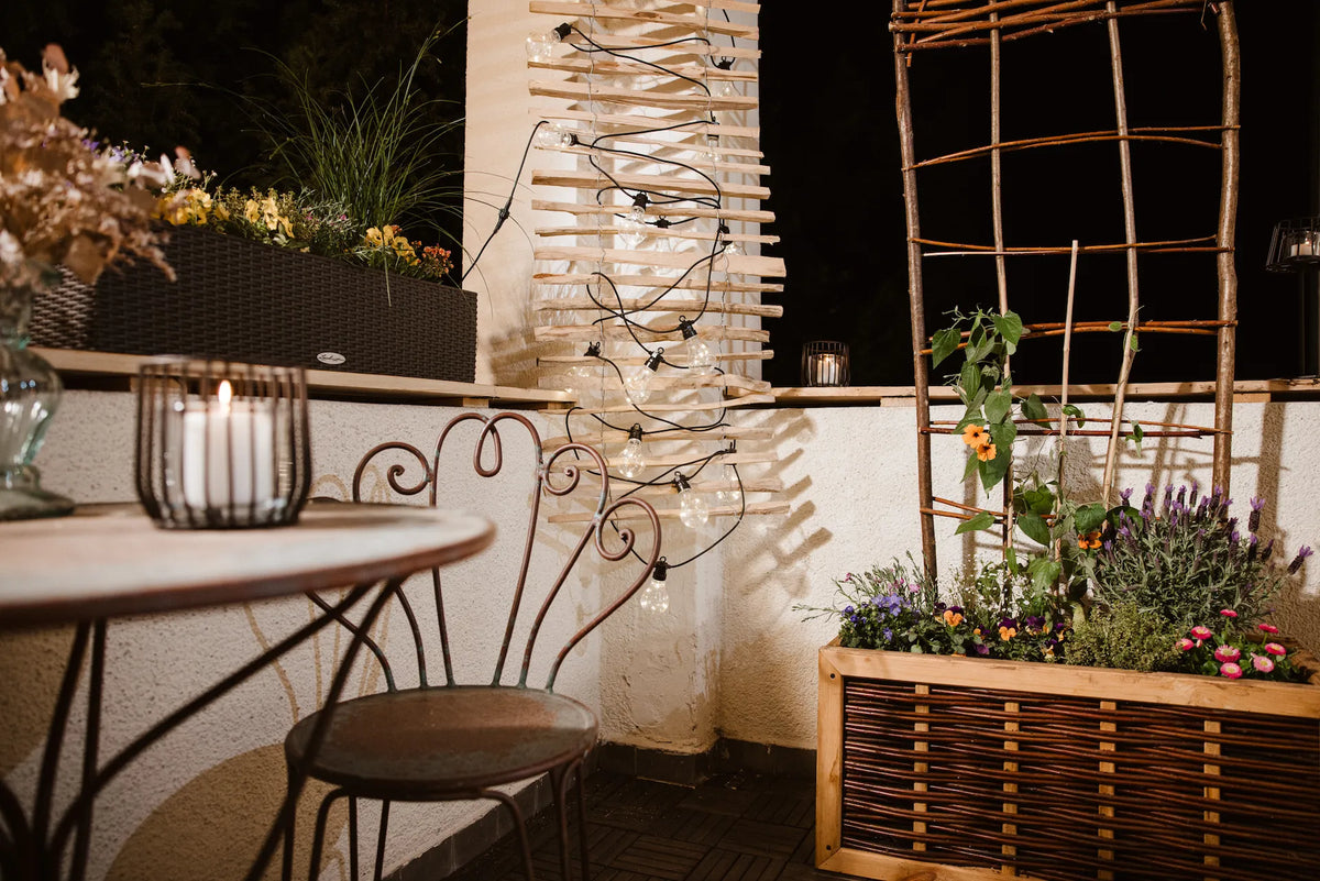 Ein stimmungsvoll beleuchteter Balkon bei Nacht mit warmen Lichterketten an einer Holzverkleidung und einer brennenden Kerze in einer Laterne auf dem Tisch. Ein bepflanzter Weiden-Blumenkasten und ein Rankgitter unterstreichen die gemütliche Wohlfühlatmosphäre.