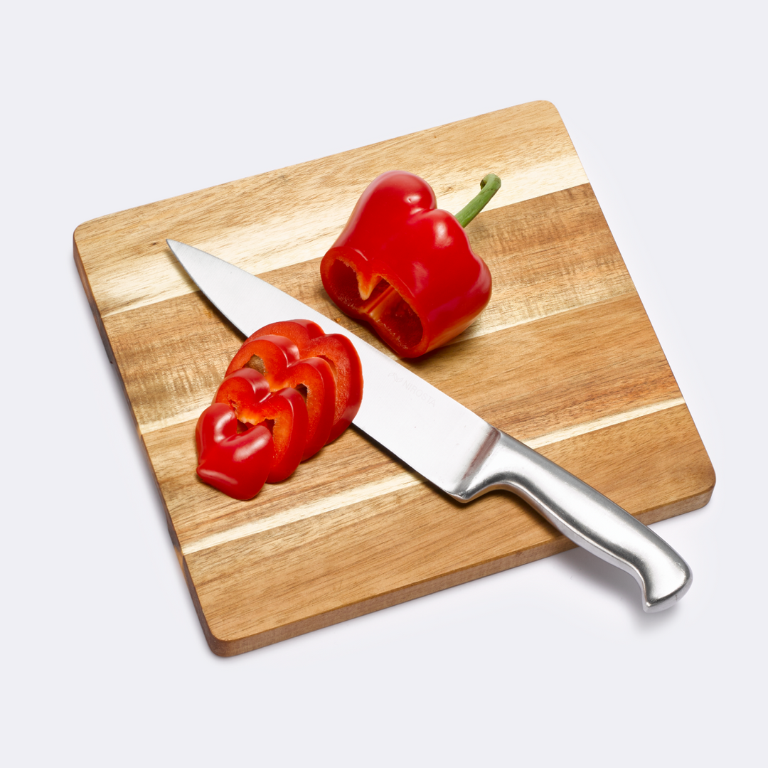 Ein Küchenmesser und eine frisch aufgeschnittene rote Paprika liegen auf einem quadratischen Schneidebrett aus hellem, klingenschonendem Holz.