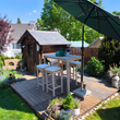 Ein kompaktes Hochtisch-Set aus hellem Akazienholz mit vier weiß gepolsterten Hockern steht auf einer Holzterrasse in einem sonnigen Garten. Im Hintergrund ergänzt ein kleines hölzernes Gartenhaus die idyllische Außenkulisse.