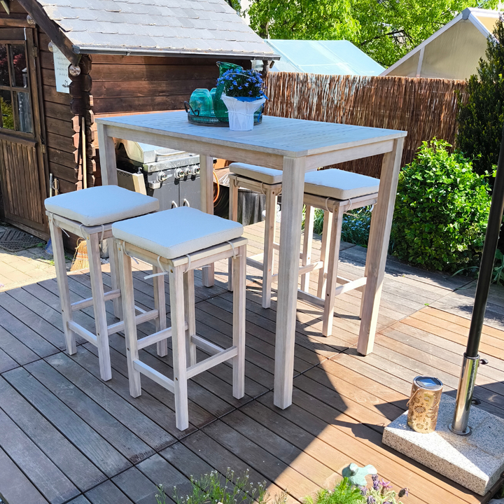 Ein kompaktes Hochtisch-Set aus hellem Akazienholz mit vier weiß gepolsterten Hockern steht auf einer Holzterrasse in einem sonnigen Garten. Im Hintergrund ergänzt ein kleines hölzernes Gartenhaus die idyllische Außenkulisse.