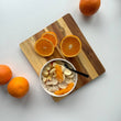 Ansicht von oben auf ein glattes Holzbrett. Darauf angerichtet sind eine Frühstücksschale mit Obst und Müsli sowie frisch aufgeschnittene Orangen.