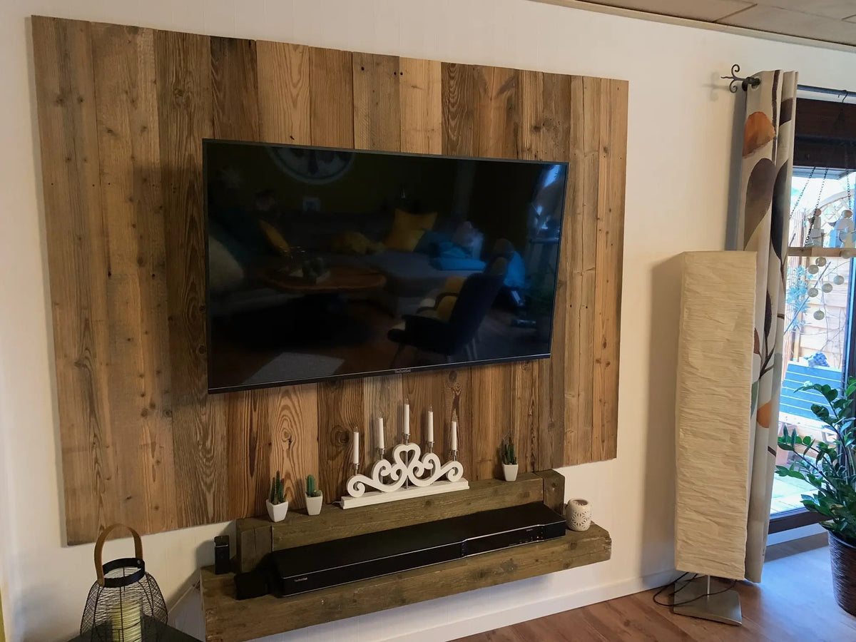 Ein Flachbildfernseher ist vor einem großflächigen Wandpanel aus vertikal angeordneten Altholz-Brettern montiert. Ein darunter hängendes, massives Holzregal bietet Platz für Dekoration wie Kerzen und kleine Kakteen, was dem Wohnzimmer eine gemütliche Atmosphäre verleiht.