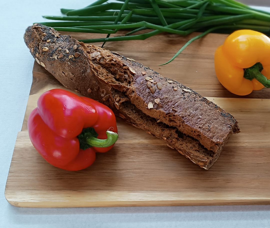 Ein dunkles Vollkorn-Baguette liegt zusammen mit einer roten und einer gelben Paprika sowie frischen Frühlingszwiebeln auf einem hochwertigen Schneidebrett aus Massivholz. 