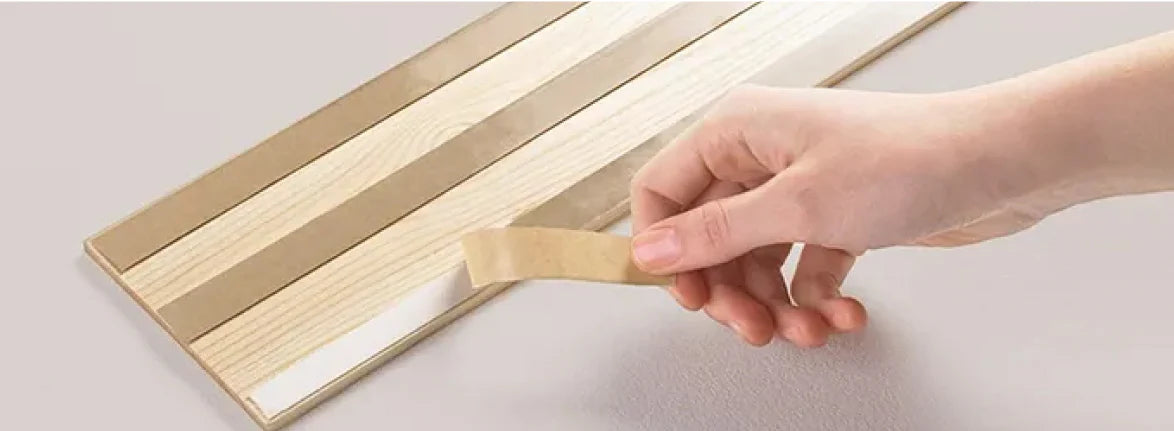Eine Hand zieht die schmale Schutzfolie von einem der rückseitig angebrachten Klebestreifen eines hellen Holzpaneels ab.