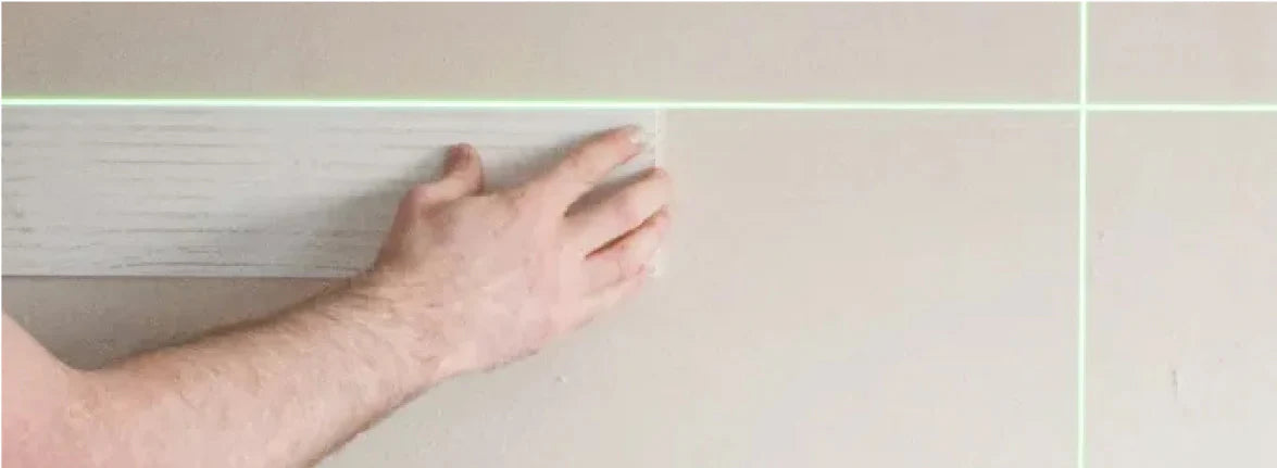 Ein hell lasiertes Holzpaneel wird mit beiden Händen fest an die Montagefläche gedrückt. Ein projiziertes grünes Laserkreuz dient dabei als optische Hilfslinie für die exakte und waagerechte Ausrichtung.