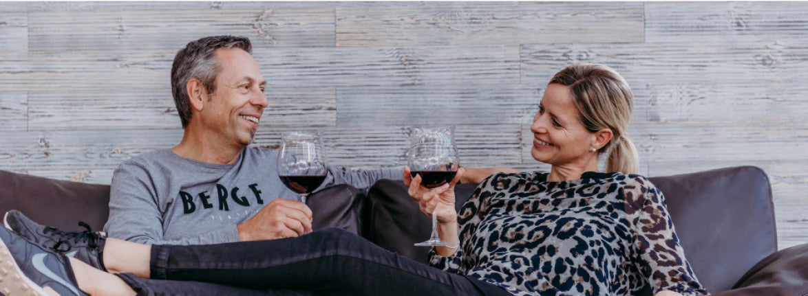 Ein Paar sitzt entspannt mit einem Glas Rotwein auf einem dunklen Ledersofa. Die Akzentwand aus grauen Holzpaneelen im Hintergrund verleiht dem Wohnbereich eine gemütliche und zugleich moderne Atmosphäre.