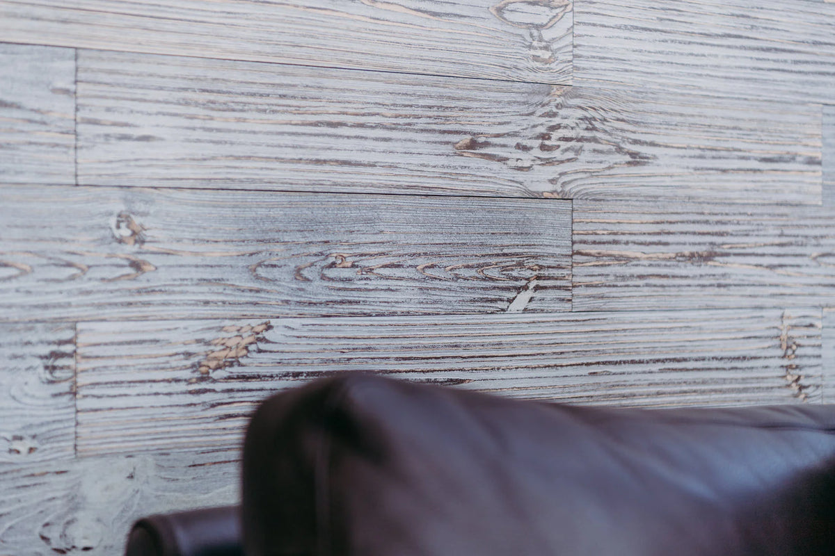 Holzpaneele mit einer grauen, strukturierten Oberflächenbehandlung, die optisch an eine Steinmauer erinnern. Im unscharfen Vordergrund ist ein dunkles Polstermöbel angeschnitten, was die Integration der Paneele in einen modernen Wohnraum veranschaulicht.